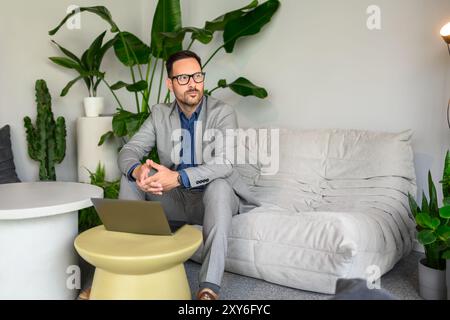 Thoughtful young businessman in eyeglasses looking away while working over laptop on sofa in office Stock Photo