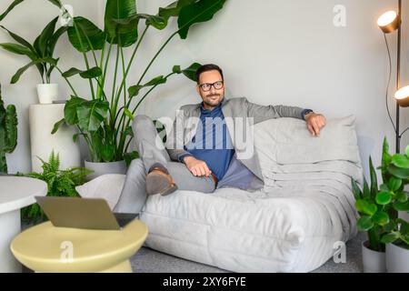 Thoughtful male CEO in eyeglasses looking away while sitting comfortably on sofa at modern workplace Stock Photo