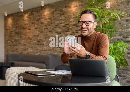 Young project manager reading e-mails over mobile phone while sitting with laptop at table in office Stock Photo