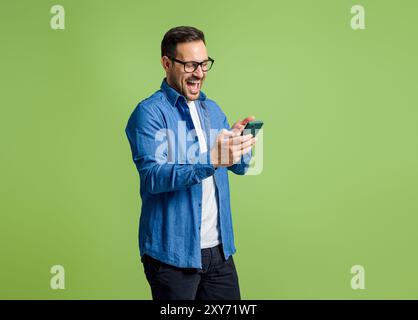 Young businessman reading good news over smart phone and shouting ecstatically on green background Stock Photo
