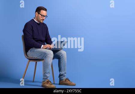 Portrait of male entrepreneur reading e-mails over laptop and sitting on chair over blue background Stock Photo
