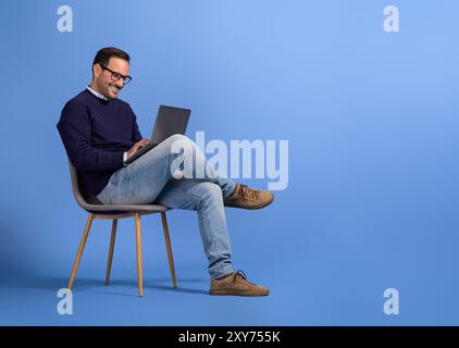 Confident young businessman in eyeglasses using laptop while sitting on chair over blue background Stock Photo