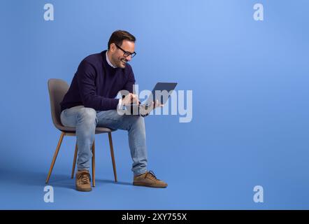 Smiling businessman checking online websites over laptop while sitting on chair over blue background Stock Photo