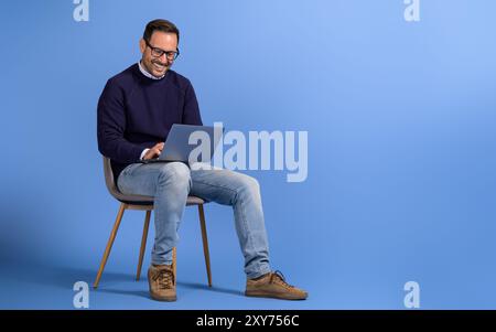 Happy male freelancer working on project over laptop while sitting on chair against blue background Stock Photo