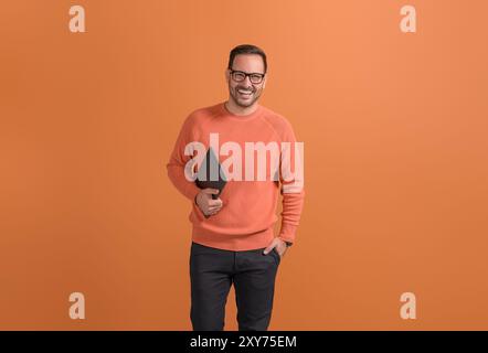 Successful happy male freelancer with hand in pocket holding laptop and posing on orange background Stock Photo