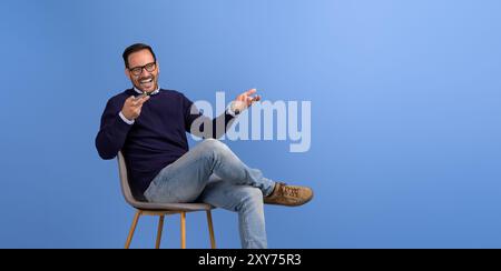 Young businessman laughing and talking on speakerphone while sitting on chair over blue background Stock Photo