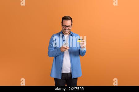 Smiling young man buying online over smart phone and paying through credit card on orange background Stock Photo