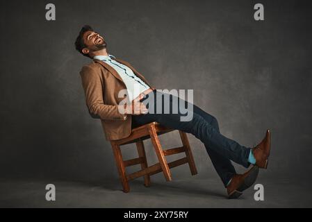 Handsome man, laughing and leaning with chair for funny humor, joke or comedy on a gray background. Young, male person or model with smile in laughter Stock Photo
