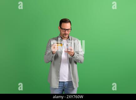 Young businessman transferring money with credit card and mobile phone on isolated green background Stock Photo