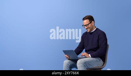 Smiling businessman doing online research over wireless computer on chair against blue background Stock Photo