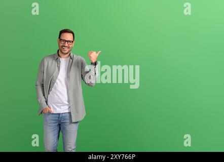 Portrait of salesman aiming thumbs at copy space and demonstrating new product on green background Stock Photo