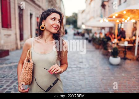 Portrait of young fashionable woman wearing summer outfit on city street by restaurant in evening. Stylish straw handbag Stock Photo