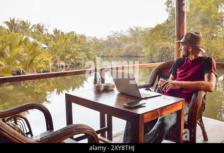 Close up thoughtful young caucasian man tourist sit by table in cafe use laptop on holidays in southeast asia in beautiful tropical nature. Remote wor Stock Photo