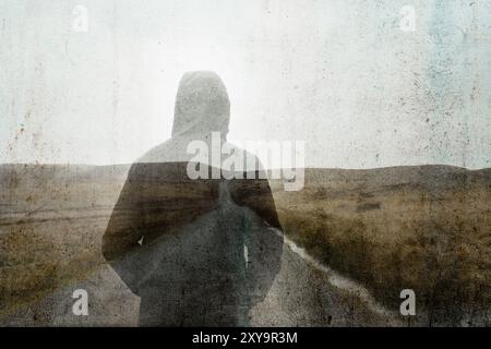A double exposure of a mysterious hooded figure, deep in thought. Standing on a bleak moorland road. With a grunge edit Stock Photo
