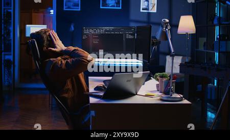 Fatigued software programmer working overnight at home, taking break from coding to stretch at desk chair. Overworked IT professional feeling exhausted, pausing tasks to rest, camera B Stock Photo