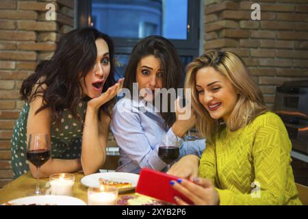 Three female friends drinking red wine in the kitchen and looking at mobile phone Stock Photo