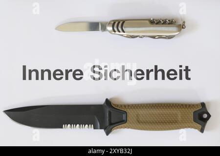 A hunting knife with a beige handle and a Swiss Army knife lie on a white surface next to a text that reads 'Homeland Security' Stock Photo