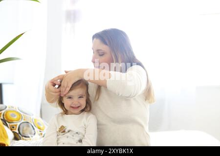 Happy loving family. Mother and baby girl having fun making hairdress sitting on the bed Stock Photo