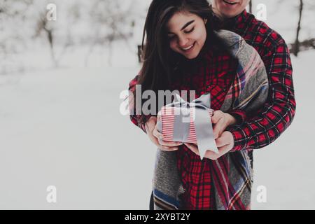 Boyfriend giving a gift box present with ribbon outdoor due to Saint Valentine day to her girlfriend. Snowy forest, trees with snow. Two are dressed i Stock Photo