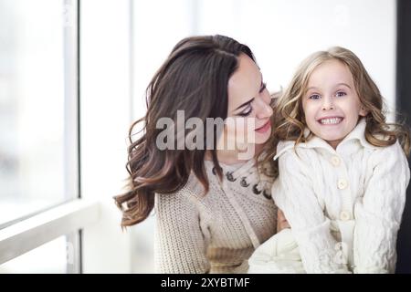 Happy loving family together. Mother and child daughter hugging by the window wearing cozy sweaters Stock Photo