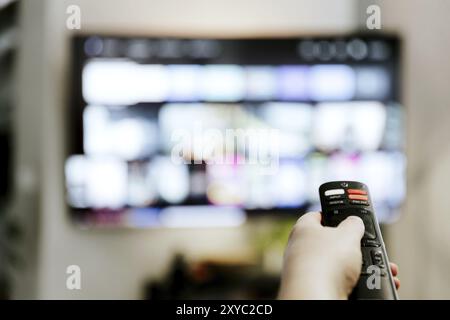 Hand holding remote control pointing to TV. Person watching smart television indoors. Blurred background. Colorful screen. Streaming series, channels. Stock Photo