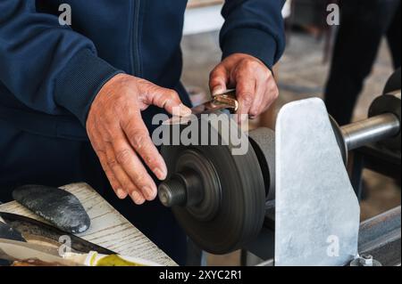 grinder sharpens the knife by holding it in hands on the abrasive rotating wheel of the sharpening machine in workshop Stock Photo