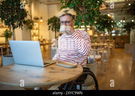Interested man with disability drinking coffee in cafe freelance working online, watching on laptop. Stock Photo