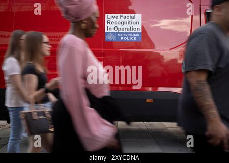Blurred shoppers walk past a Met Police van that's using Artificial Intelligence (AI) Facial Recognition (FI) technology to spot persons of interest, and pre-empt criminal activity, on Oxford Street, on 28th August 2024, in London, England. Facial Recognition (FR) technology is used by the Met to prevent and detect crime, find wanted criminals, safeguard vulnerable people. Real time surveillance helps officers locate people on a ‘watchlist’ who are sought by the police. FR cameras focus specific areas and  when people pass through, their images are streamed directly to the Live Facial Recognit Stock Photo