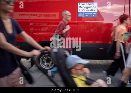 Blurred shoppers walk past a Met Police van that's using Artificial Intelligence (AI) Facial Recognition (FI) technology to spot persons of interest, and pre-empt criminal activity, on Oxford Street, on 28th August 2024, in London, England. Facial Recognition (FR) technology is used by the Met to prevent and detect crime, find wanted criminals, safeguard vulnerable people. Real time surveillance helps officers locate people on a ‘watchlist’ who are sought by the police. FR cameras focus specific areas and  when people pass through, their images are streamed directly to the Live Facial Recognit Stock Photo