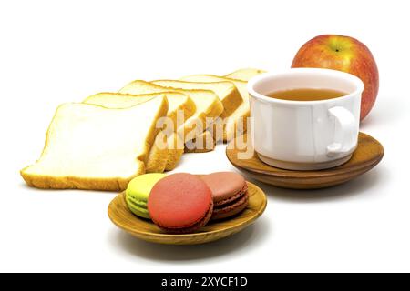 Colorful macarons on wooden plate, sliced bread, red apple and a cup of hot tea on white background Stock Photo