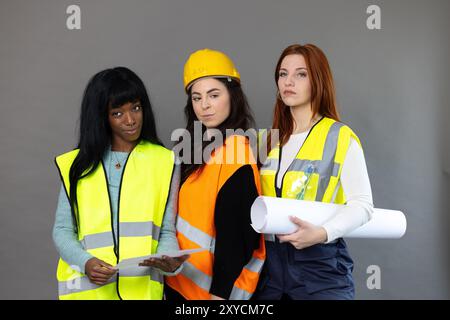 Three young female engineers wearing safety vests and holding blueprints are standing in front of a grey background Stock Photo