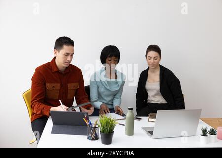 Three focused colleagues are collaborating on a project, using digital tablets and laptops at a shared desk Stock Photo