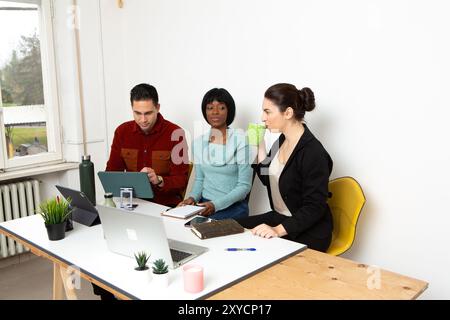 Three young professionals are sitting at a desk, working on a project together and using laptops and tablets Stock Photo