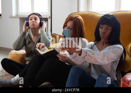 Three female friends are spending their free time together at home, each absorbed in her favorite activity Stock Photo