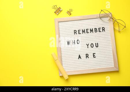 Letter board with phrase Remember Who You Are, glasses, marker and binder clips on yellow background, flat lay. Space for text Stock Photo