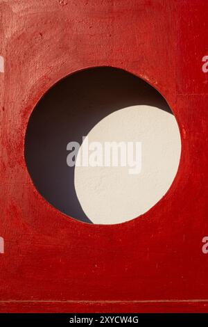 Detail of a  bright red case with a circle hole, prepared for a fire extinguisher. Empty in this case. Sunlight and shadow creating a shape of moon. D Stock Photo