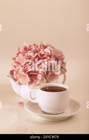 Porcelain teapot and cup fo the tea with bunch of petals of pink flowers placed against beige background Stock Photo