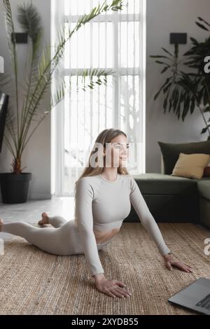 Healthy sport lifestyle concept, happy smiling blogger girl doing yoga exercises at home with green loft interior watching and repeating trainer exerc Stock Photo