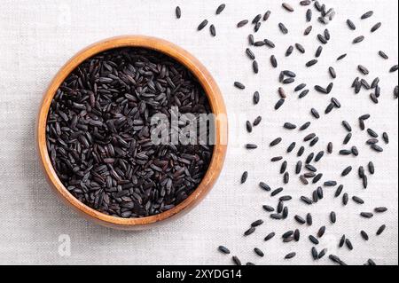 Black rice, purple or forbidden rice in a wooden bowl on linen fabric. Oryza sativa, Asian cultivated rice, cereal grain with deep black color. Stock Photo