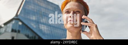 Woman with red hair smiles, talks on phone with campus building in background Stock Photo
