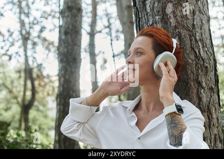 A young teacher with red hair listens to music with headphones while leaning against a tree. Stock Photo