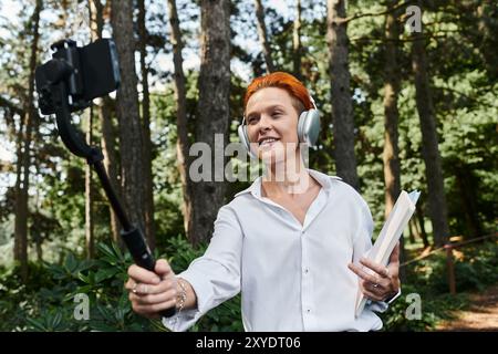 A teacher with red hair takes a selfie with a selfie stick while walking through a forest. Stock Photo