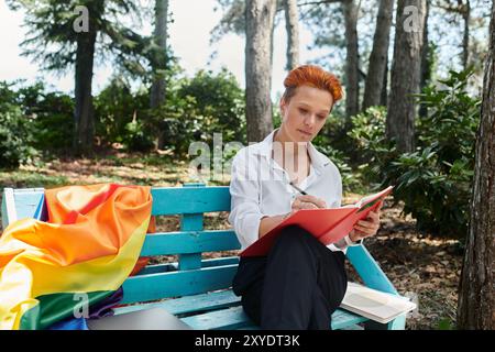 A woman in a white shirt sits on a bench in a campus grove, writing in a red notebook. Stock Photo