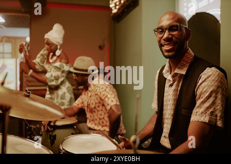 Young African American man sitting at drum kit and smiling at camera while two musicians getting ready for performance in background Stock Photo