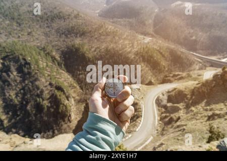 Close-up A man's hand holds a pocket compass against the backdrop of a mountain road and forest. The concept of outdoor navigation Stock Photo