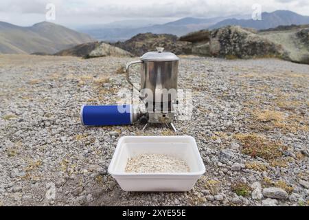 A plastic cup of oatmeal waiting for a while in a metal mug on a portable torch with a gas cylinder will boil water. The concept of food in the campai Stock Photo