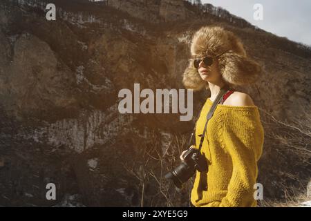 A girl photographer in sunglasses and a big fur hat and a yellow knitted sweater stands against the background of high rocks in the gorge with a camer Stock Photo