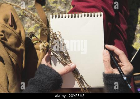 Close-up on the hands of a girl holding a blank notebook. A dry bouquet of herbs in her hand and a pencil. Travel designer artist Stock Photo