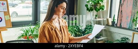 A woman in a beige blouse reviews papers, standing in a modern office with large windows and greenery. Stock Photo