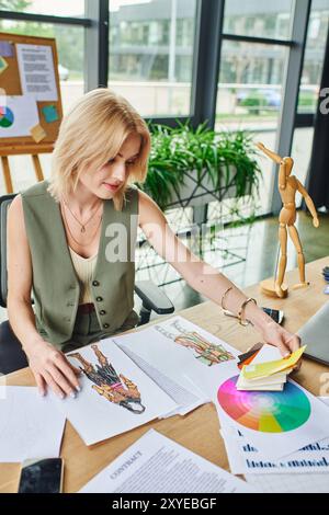 A blonde woman in a green vest and white top, sitting at her desk, reviewing fashion sketches and color swatches. Stock Photo